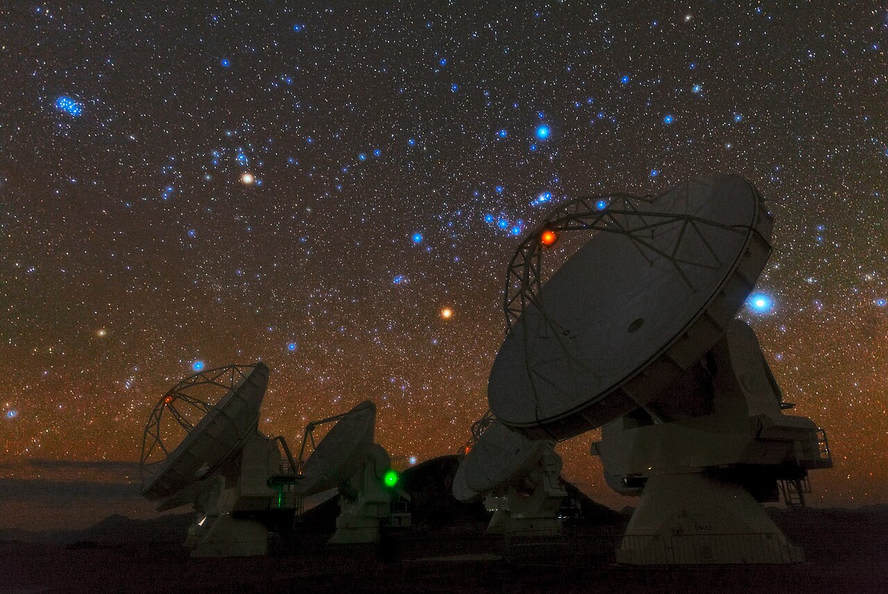 Snímek ukazující noční hvězdnou jižní oblohu nad talířovými anténami radioteleskopu ALMA. Vpravo je jasná hvězda Sirius, uprostřed souhvězdí Orionu, vlevo souhvězdí Býka a u levého okraje Plejády.